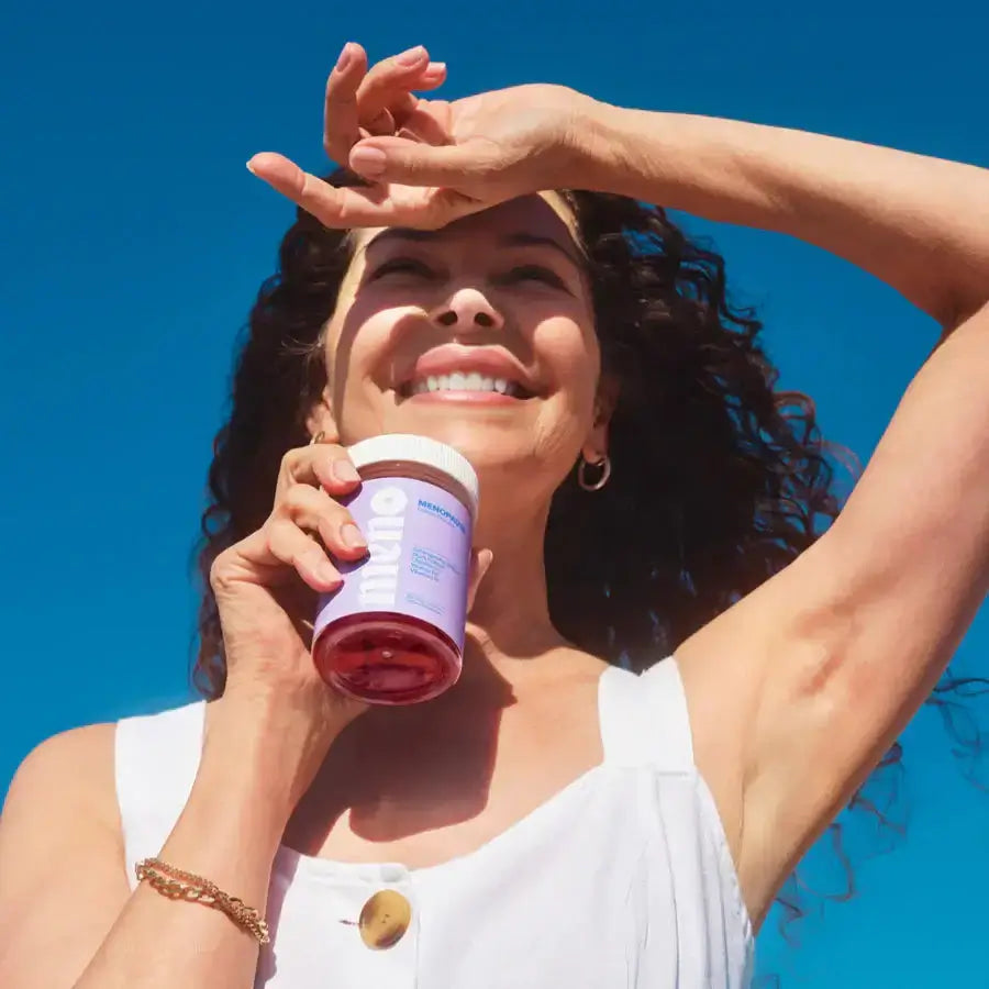 Smiling woman holding a supplement jar under a clear blue sky, enjoying the sunny day.